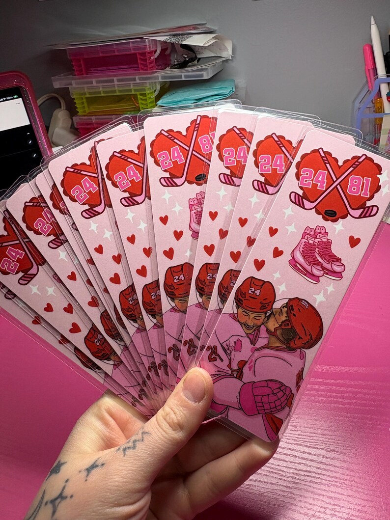 May include: A stack of pink hockey-themed cards featuring a heart-shaped design with crossed hockey sticks, the number 24, and a pair of skates. The cards also depict two hockey players embracing. Small red hearts and stars decorate the cards.