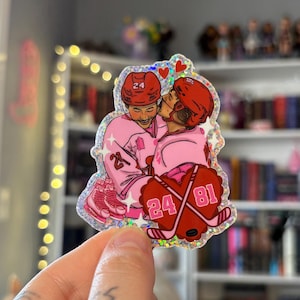 May include: A holographic sticker featuring a cartoon illustration of two hockey players in pink uniforms embracing. The players wear red helmets with the number 24. Crossed hockey sticks with the numbers 24 and 81 are also shown.