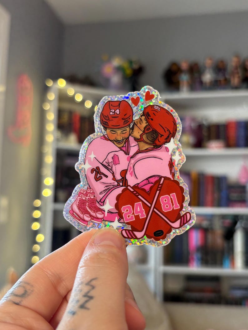 May include: A holographic sticker featuring a cartoon illustration of two hockey players in pink uniforms embracing. The players wear red helmets with the number 24. Crossed hockey sticks with the numbers 24 and 81 are also shown.