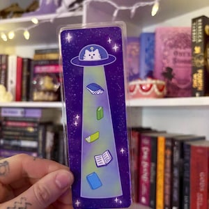 May include: A whimsical bookmark featuring a cat in a UFO beaming up books. The background is a starry purple sky. The bookmark is rectangular and made of clear plastic.