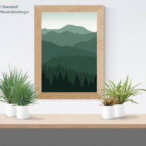 Op de afbeelding: Een digitale download van een printbare kunstprint met een minimalistische illustratie van een bergketen met een bos op de voorgrond. De afbeelding is ingelijst en wordt weergegeven op een plank met twee planten in potten.