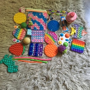 May include: A collection of colorful pop-it fidget toys, including a rainbow pop-it, a strawberry pop-it, a unicorn pop-it, a butterfly pop-it, a fish pop-it, a square pop-it, a round pop-it, a heart pop-it, a star pop-it, and a fidget spinner.