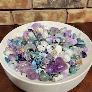 May include: A white ceramic bowl filled with an assortment of polished gemstones and small glass bottles with cork stoppers. The gemstones are various shades of purple, blue, green, and white. The background features a brick wall.