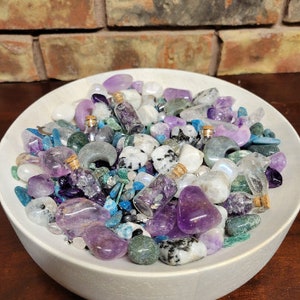 May include: A white ceramic bowl filled with an assortment of crystals and small glass bottles. The crystals are various shades of purple, white, blue, and green. The bottles have cork stoppers and contain purple crystals. The background features a brick wall.