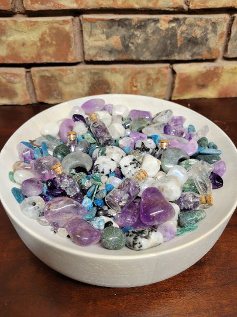 May include: A white ceramic bowl filled with an assortment of crystals and small glass bottles. The crystals are various shades of purple, white, blue, and green. The bottles have cork stoppers and contain purple crystals. The background features a brick wall.