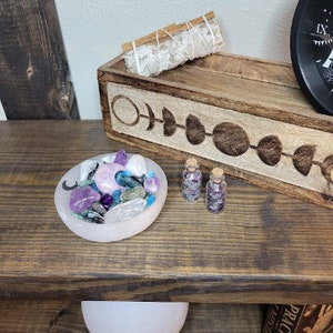 May include: A wooden shelf displays a collection of spiritual items. A pink bowl holds crystals and a crescent moon. Two small glass bottles with corks are filled with crystals. A wooden tray with moon phase carvings holds a sage bundle.
