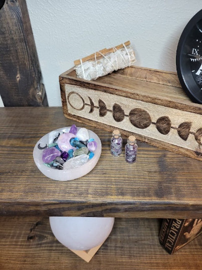 May include: A wooden shelf displays a collection of spiritual items. A pink bowl holds crystals and a crescent moon. Two small glass bottles with corks are filled with crystals. A wooden tray with moon phase carvings holds a sage bundle.