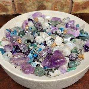 May include: A white ceramic bowl filled with an assortment of colorful gemstones and small glass bottles. The gemstones are in shades of purple, blue, green, and white. The small bottles have cork stoppers and contain purple crystals.