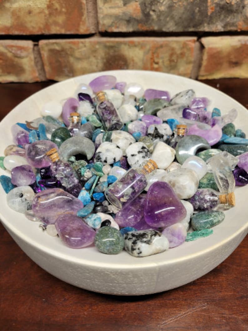May include: A white ceramic bowl filled with an assortment of colorful gemstones and small glass bottles. The gemstones are in shades of purple, blue, green, and white. The small bottles have cork stoppers and contain purple crystals.