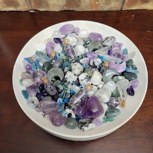 May include: A white ceramic bowl filled with an assortment of crystals and small glass bottles. The crystals are various shades of purple, blue, green, and white. The bottles contain small crystals and are sealed with corks. The bowl sits on a dark wooden surface.