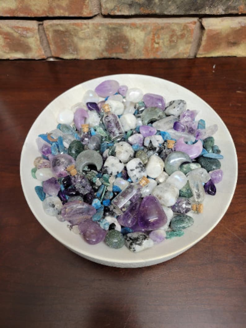 May include: A white ceramic bowl filled with an assortment of crystals and small glass bottles. The crystals are various shades of purple, blue, green, and white. The bottles contain small crystals and are sealed with corks. The bowl sits on a dark wooden surface.
