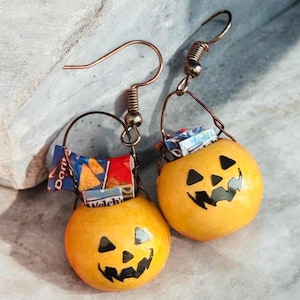 May include: A pair of dangle earrings featuring miniature orange jack-o'-lantern buckets with black painted facial features. Each bucket contains small, colorful snack bag replicas, including a Doritos bag, suspended from copper-toned metal hooks.