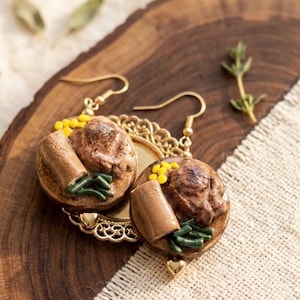 May include: A pair of dangle earrings featuring miniature food charms shaped like a roast dinner with brown meat, green beans, and yellow corn kernels. The charms are mounted on gold-toned filigree settings with gold-toned ear wires.