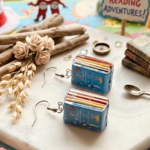May include: A pair of dangle earrings featuring miniature book charms with blue covers and multi-colored pages. Each charm is attached to a silver-toned metal fishhook ear wire. The book covers display small illustrations and text in a blue color scheme.