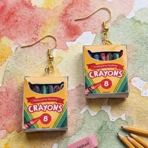 May include: A pair of dangle earrings featuring miniature yellow boxes of eight crayons. Each box displays the text Preferred by Teachers, CRAYONS, and the number 8, with a red crayon illustration. The earrings have gold-toned metal hooks.