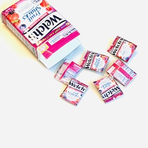 May include: A Welch's Fruit Snacks box with a pink and white design featuring images of berries and cherries, accompanied by five individual snack pouches scattered around it. The packaging displays the brand name and fruit imagery.