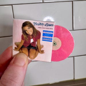 Peut inclure: Un disque vinyle rose avec un label blanc et un motif de fleur verte. Le disque est dans une pochette en plastique transparente avec une couverture représentant une photo de Britney Spears et le texte "...baby one more time" et "Édition limitée".