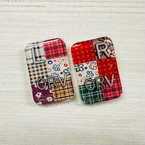 May include: Two rectangular acrylic charms featuring a patchwork design of plaid and floral patterns in red, white, blue, and green. The left charm displays the silver letters LORV, while the right charm displays the silver letters RORV.