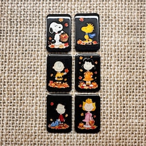 May include: A set of six rectangular black tiles featuring Peanuts characters in autumn settings. The tiles display Snoopy with a basket, Woodstock, Charlie Brown, Lucy, Linus, and Sally, all surrounded by colorful fall leaves and pumpkins.