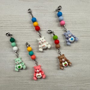 May include: Five decorative charms featuring miniature bear figures with colorful beads attached to silver-toned lobster clasps. The bears include a mint green clover bear, a pink rainbow bear, a white rainbow bear, a brown strawberry bear, and a blue cloud bear.