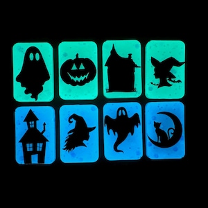 May include: Eight rectangular glow-in-the-dark tiles featuring black silhouettes of Halloween icons including ghosts, a jack-o'-lantern, haunted houses, a witch, and a cat on a crescent moon. The tiles emit a bright cyan light against a black background.