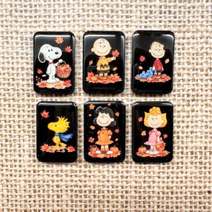 May include: Six rectangular black cabochons featuring Peanuts characters in autumn settings. Snoopy holds a basket of apples, Charlie Brown stands in leaves, Linus holds a blanket, Woodstock wears a scarf, Lucy stands by pumpkins, and Sally holds a basket.