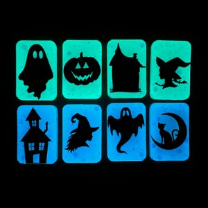 May include: Eight rectangular glow-in-the-dark items arranged in two rows. The top row features black silhouettes of a ghost, a jack-o'-lantern, a house, and a witch. The bottom row displays a house, a witch profile, a ghost, and a cat on a moon.