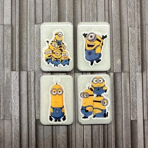 May include: Four rectangular light gray magnets featuring yellow Minion characters. The top left shows a group of five Minions, the top right a single cheering Minion, the bottom left a tall single Minion, and the bottom right three stacked Minions.