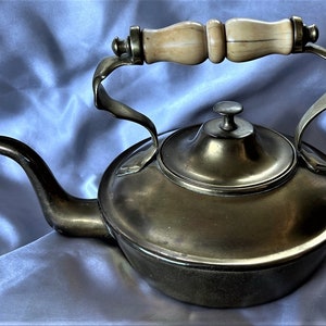Pode incluir: Bule de chá de latão antigo com um bico curvo e uma alça com uma pega de madeira clara. O bule tem um corpo redondo e uma tampa com um pequeno botão. A alça é fixada ao corpo com suportes de metal curvos.