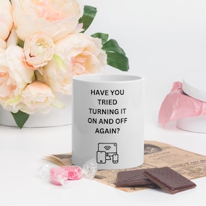 Op de afbeelding: Witte keramische mok met de tekst "HAVE YOU TRIED TURNING IT ON AND OFF AGAIN?" en een afbeelding van een computer, telefoon en tablet. De mok staat naast een boeket roze bloemen en twee chocoladerepen.