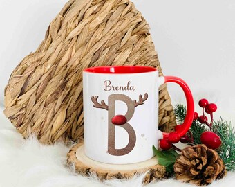 May include: White ceramic mug with a red rim and handle. The mug features a brown glitter letter "B" with a reindeer face on top. The name "Brenda" is printed above the letter. The mug is sitting on a wooden coaster.