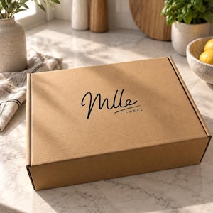 May include: A rectangular brown cardboard shipping box with a hinged lid. The top of the box features the black printed text Mlle LABEL in a stylized script font. The box is constructed from corrugated paper material with a smooth finish.