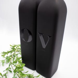 May include: Two black glass bottles with the letters "O" and "V" embossed on the front. The bottles are sitting on a white surface.