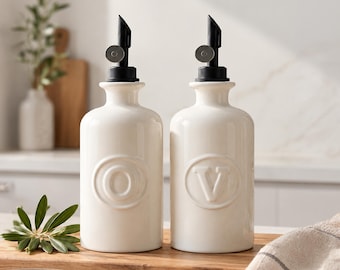 May include: Two white ceramic oil and vinegar bottles with black pour spouts. Each bottle features a raised circular emblem on the front, one embossed with the letter O and the other with the letter V, standing on a wooden surface.
