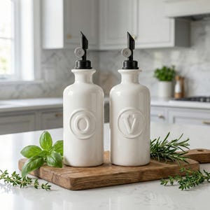 Peut inclure: Deux bouteilles blanches en céramique pour huile et vinaigre, avec des becs noirs et des lettres en relief "O" et "V". Les bouteilles sont posées sur une planche à découper en bois avec des herbes fraîches.