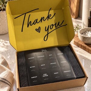 May include: A cardboard gift box with a yellow interior lid featuring the black text Thank you and a heart icon. Inside, a black tray holds twelve small square packets labeled with herbs and spices including Lavender, Marjoram, Mint, Mustard, Nutmeg, Onion, Oregano, Paprika, Parsley, Pink Pepper, Poppy, and Provence.