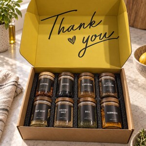 May include: A cardboard gift box with a yellow interior lid featuring the black text Thank you and a heart icon. Inside are eight glass spice jars with wooden lids and black labels for Cayenne, Sesame, Cinnamon, Steak Seasoning, Bay Leaf, Celery Salt, Dill, and Turmeric.