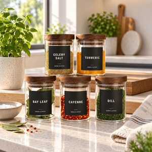 May include: Five clear glass spice jars with dark wooden lids, each featuring a black square label with white text reading Celery Salt, Turmeric, Bay Leaf, Cayenne, or Dill, and the subtext Herbs & Spices, arranged on a kitchen counter.