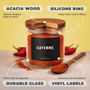 May include: A clear glass spice jar filled with red cayenne pepper, featuring an acacia wood lid with a silicone ring. The jar has a black vinyl label with the text CAYENNE HERBS & SPICES. A wooden spoon rests nearby with loose pepper.