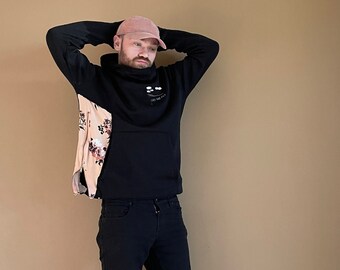 May include: Black hoodie with a floral pattern on the side and a pink baseball cap. The hoodie has the text "Les Nouveaux" on the front. The person is wearing black jeans.