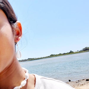 May include: A white shell necklace and matching earrings are displayed against a bright blue sky and water. The jewellery is made of white shells strung together, creating a coastal, beachy aesthetic.