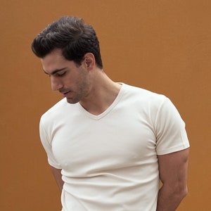 以下が含まれることがあります： 茶色の壁の前に立っている男性。白いVネックの半袖Tシャツを着ています。