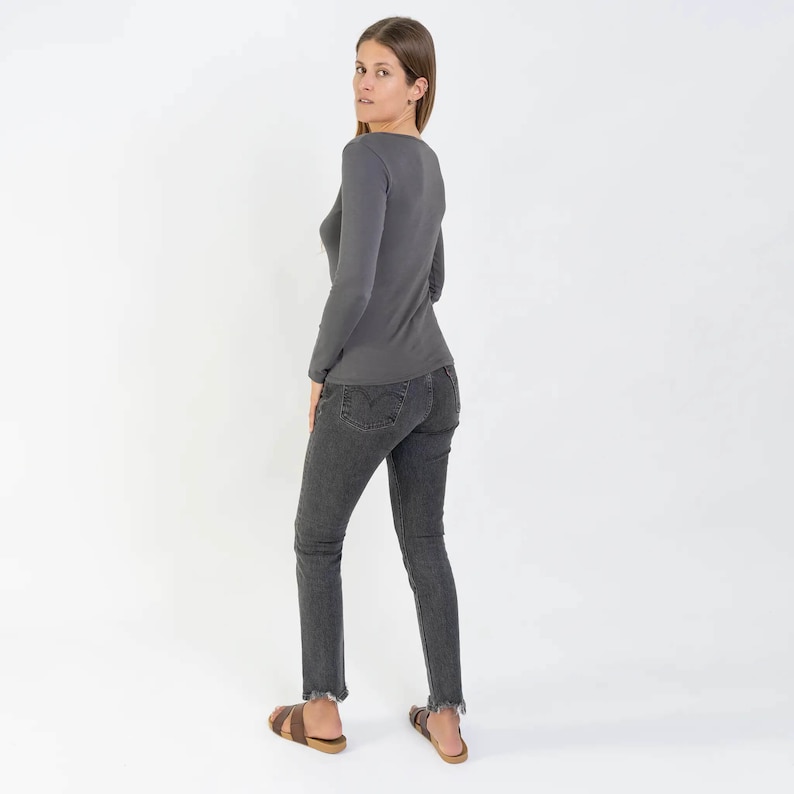 May include: A woman wearing a gray long-sleeved shirt and dark wash jeans. She is standing with her back to the camera and her hair is pulled back.