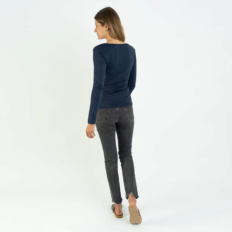 May include: A navy blue long-sleeved shirt with a scoop neck. The shirt is fitted and has a slightly cropped length. The model is wearing dark wash jeans and brown sandals.