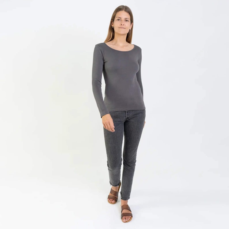 May include: A woman wearing a charcoal gray long-sleeved shirt with a scoop neck and dark gray jeans. She is wearing brown sandals.