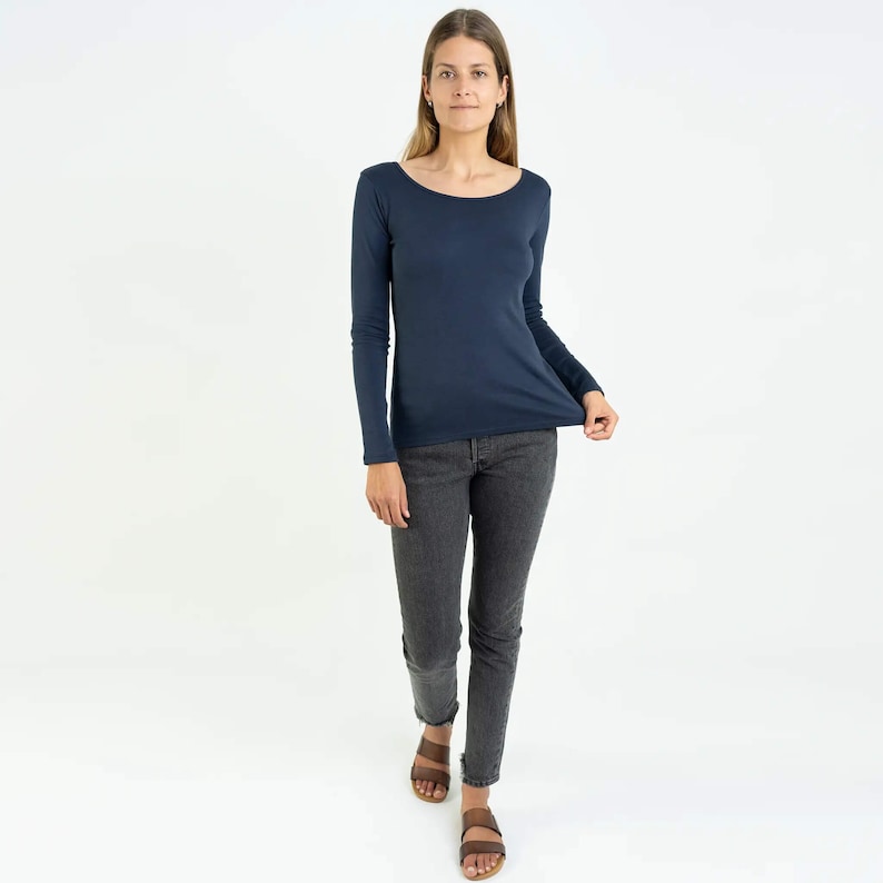 May include: A woman wearing a navy blue long-sleeved shirt and dark gray jeans. She is standing with her feet slightly apart and her hands at her sides.