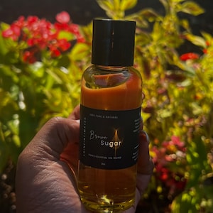 May include: A clear glass bottle with a black cap, filled with a golden-brown liquid. The label reads "Brown Sugar" and "Yoni Essential Oil Blend". The bottle is held in a hand, with a blurred background of green and red foliage.