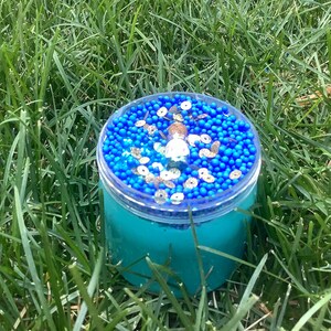 May include: A clear plastic container filled with light blue slime. The slime is topped with blue and gold beads.
