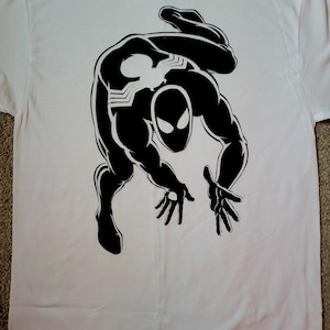 K&ouml;nnte beinhalten: Wei&szlig;es T-Shirt mit einer schwarzen Grafik eines Superhelden in dynamischer Pose. Der Superheld hat ein spinnenartiges Emblem auf Brust und R&uuml;cken und eine Maske mit gro&szlig;en, ausdrucksstarken Augen. Comic-Design.