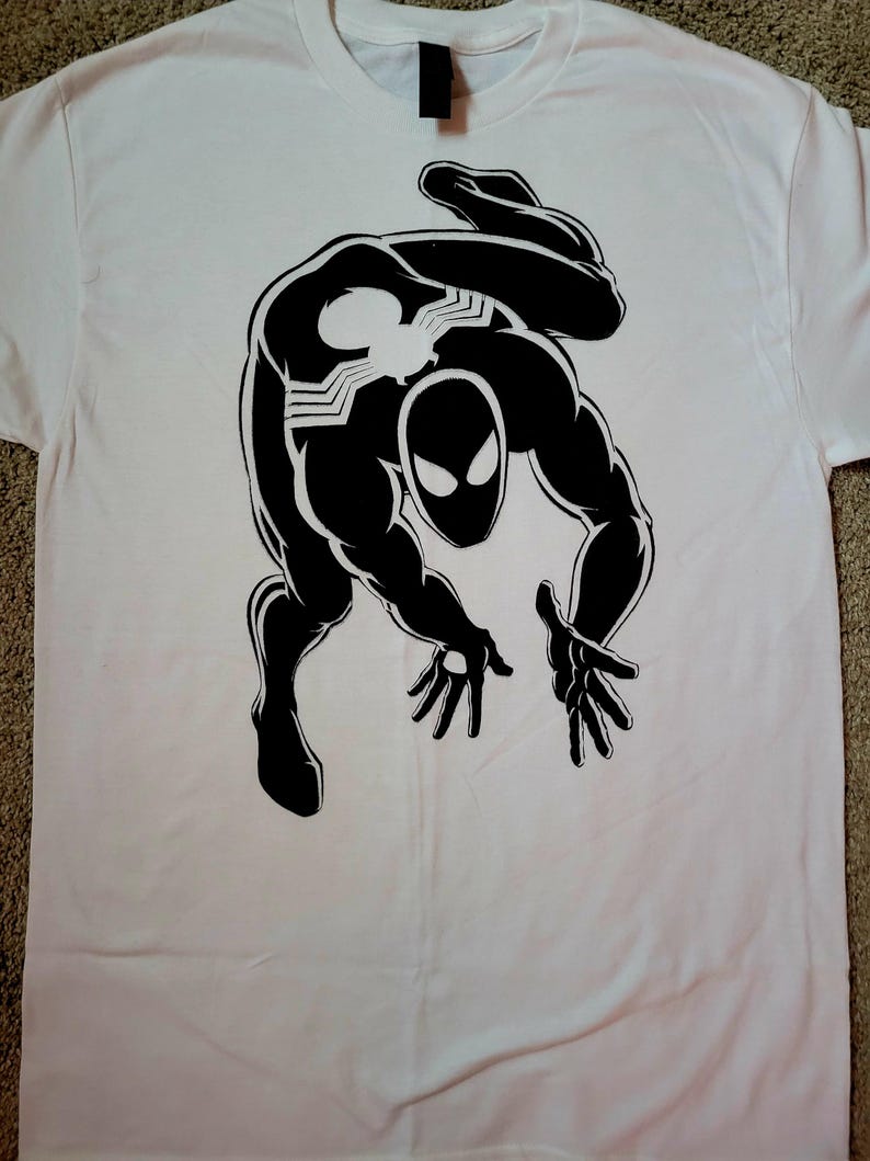 K&ouml;nnte beinhalten: Wei&szlig;es T-Shirt mit einer schwarzen Grafik eines Superhelden in dynamischer Pose. Der Superheld hat ein spinnenartiges Emblem auf Brust und R&uuml;cken und eine Maske mit gro&szlig;en, ausdrucksstarken Augen. Comic-Design.